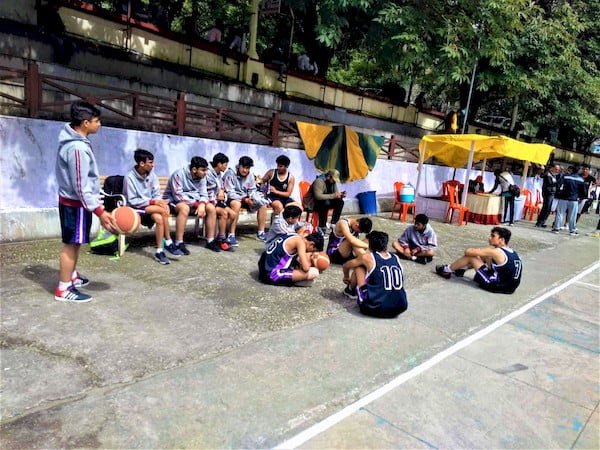 One Basketball player standing with an orange Basketball in his hand, five sitting on the ground with an orange basketball & five sitting on benches in blue dresses & two red lined hoody jaickets discussing about the game before their practice.