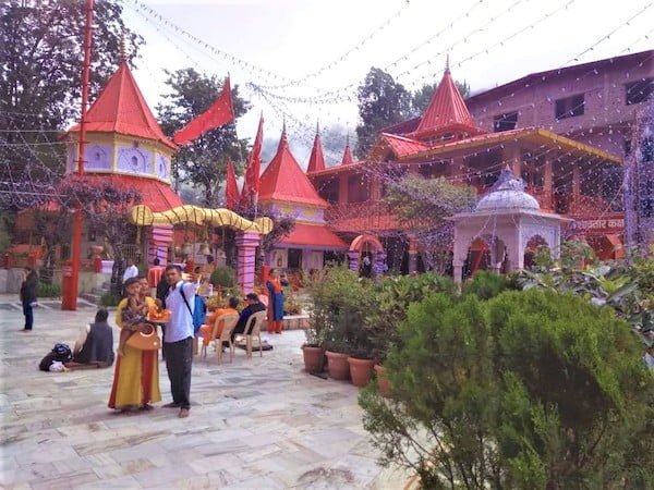 A man in sky blue shirt & black paint carrying a backpack taking selfie photo with his wife dressed in yellow suit carrying pooja thali, golden coloured handbag & her small kid in safron coloured Naina Devi mandir complex among many sitting & standing devotees in front of green decorative small plants.