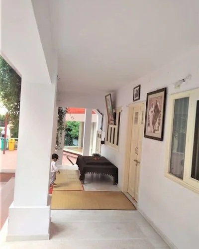 A small kid walking towards black blancket covered Chowki of Baba Neem Karoli in the gallery of a white coloured building with L-shaped cream cloured doormat near the chowkiand three well framed photographs hanging on the wall. 