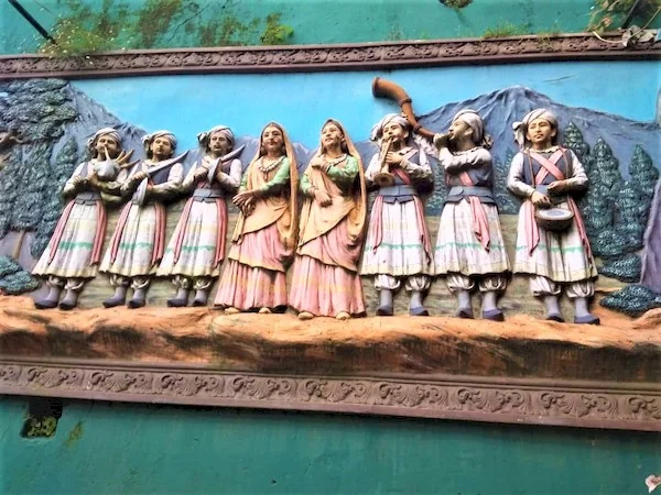 Wall Sculpture of two ladies singers in the middle, three male artists on the left & three on the right Performing Artists in traditional dresses with blue sky, mountains & Forest bacdrop.