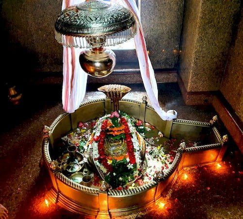 White Dhoti knot to the outer wall of Shri Mahadev Ji Shiva Linga from upside & 5 burning candles on the front side outer wall at Garbha Griha. Om written Chatri with Lota hung over flowers decorated Shiva Linga covered by a snake with pooja samagri & Utensils.